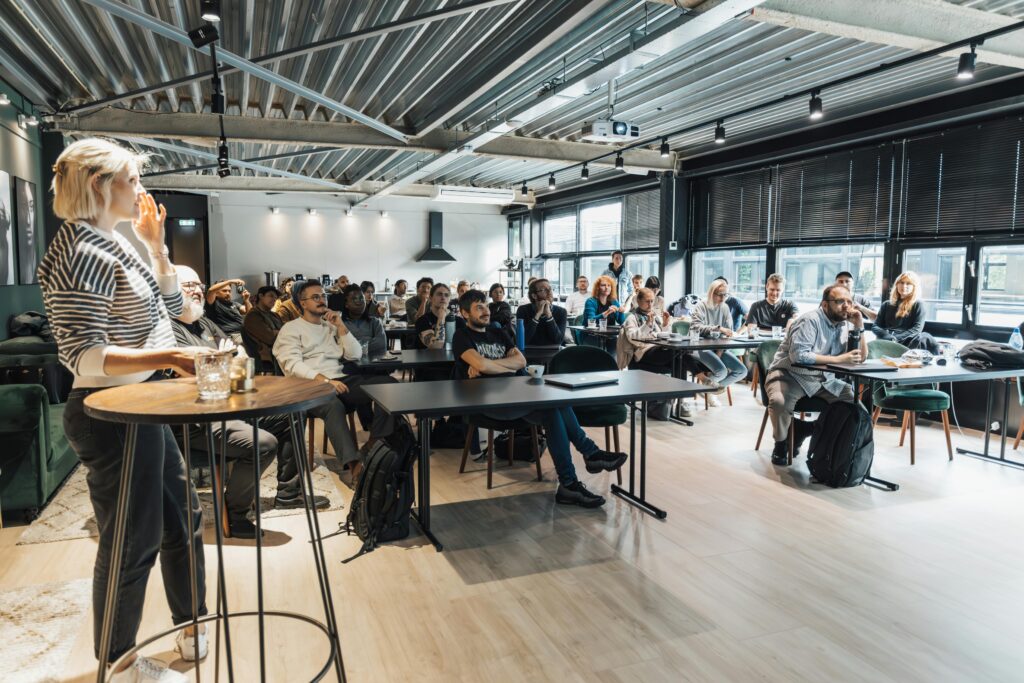 A woman standing at a high table in the foreground speaks to a large, attentive audience seated in a modern, industrial-style room with exposed ceiling beams. 